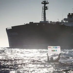 Eine Person – im Wasser schwimmend – hält ein Banner in die Höhe auf dem steht: „Oil Fuels War“. Im Hintergrund sieht man einen riesigen russischen Öltanker