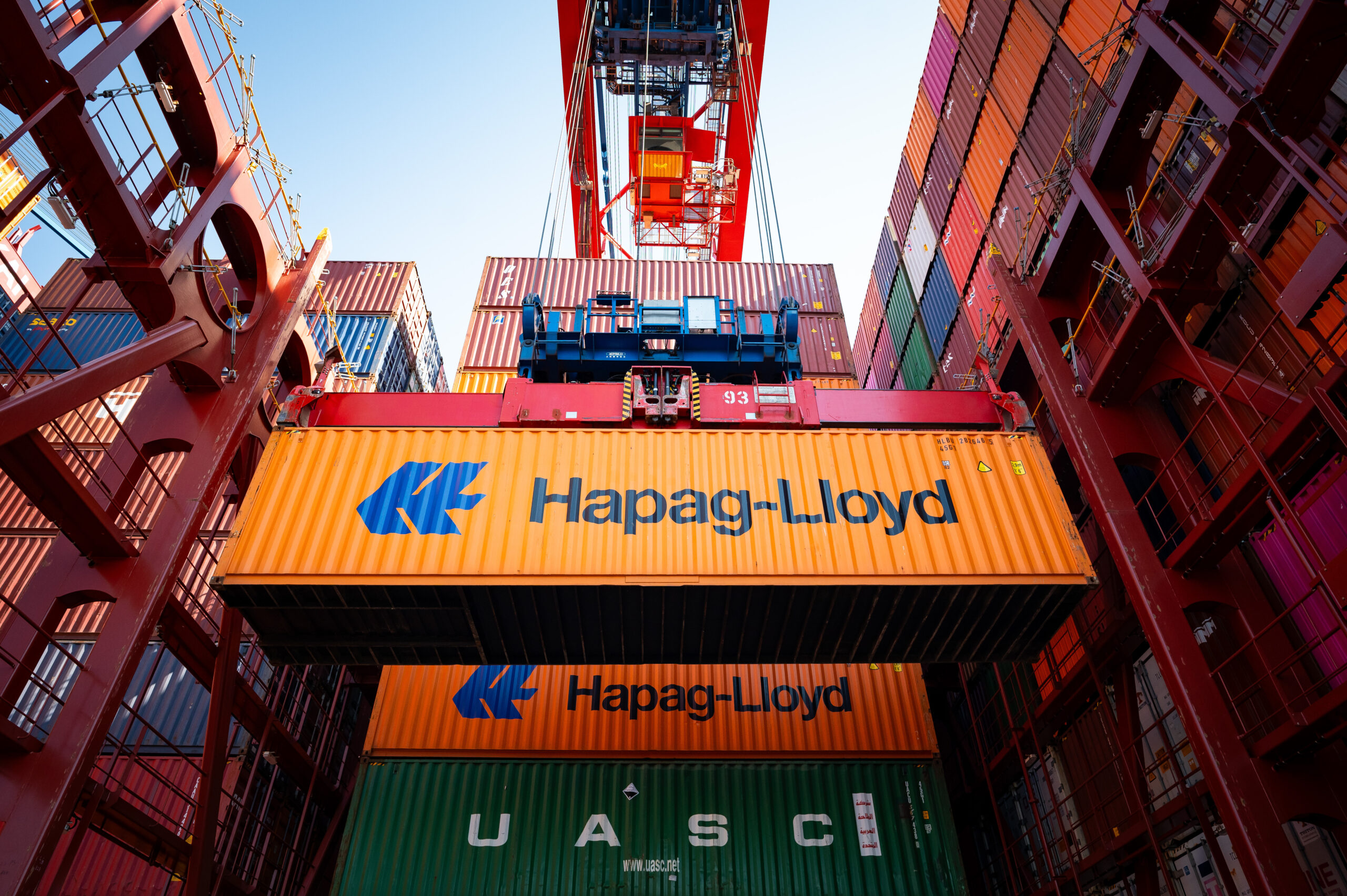 Ein Container von Hapag Lloyd hängt an einem Krahn. Hinter ihm, dutzende Container mehr. Ein Frachtschiff wird beladen.