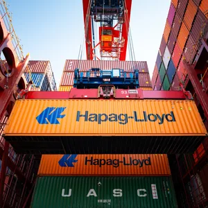 Ein Container von Hapag Lloyd hängt an einem Krahn. Hinter ihm, dutzende Container mehr. Ein Frachtschiff wird beladen.