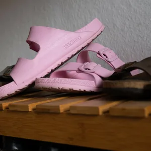 Mehrere Paare der Sandalen Birkenstocks liegen auf einer Holzbank. In der Mitte ist ein Pinkes Paar, auf dem der Name der Firma zu lesen ist.