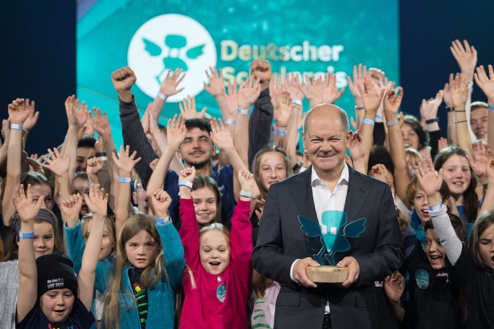 Olaf Scholz hält den Deutschen Schulpreis in der Hand. Im Hintergrund stehen jubelnde Kinder mit erhobenen Händen.