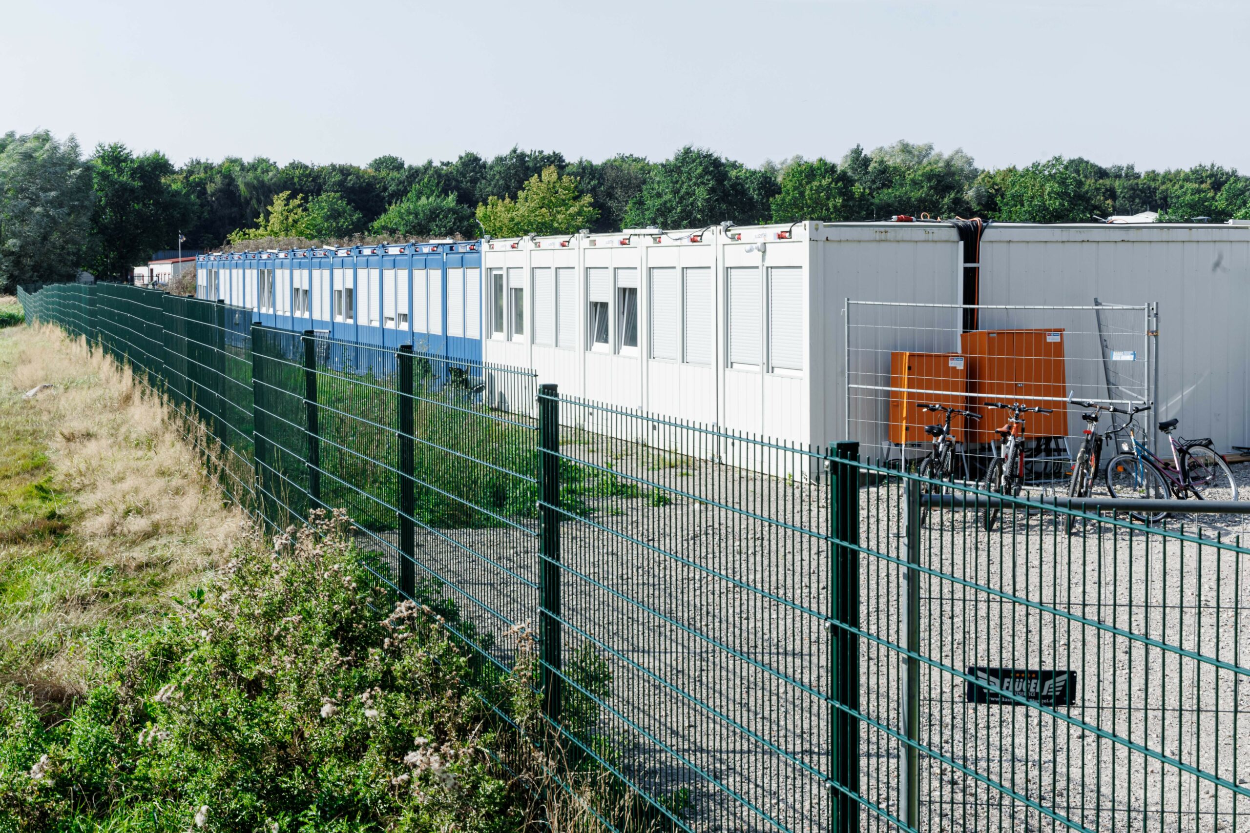 Container als Unterkunft von geflüchteten Menschen stehen aneinandergereiht hinter einem Zaun