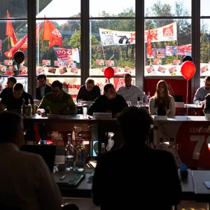 Vor einem großen Fenster, wo draußen viele Fahnen und Schilder einer Demonstration zu sehen sind, steht ein langer Tisch in einem dunklen Raum an dem viele Menschen sitzen und verhandeln.