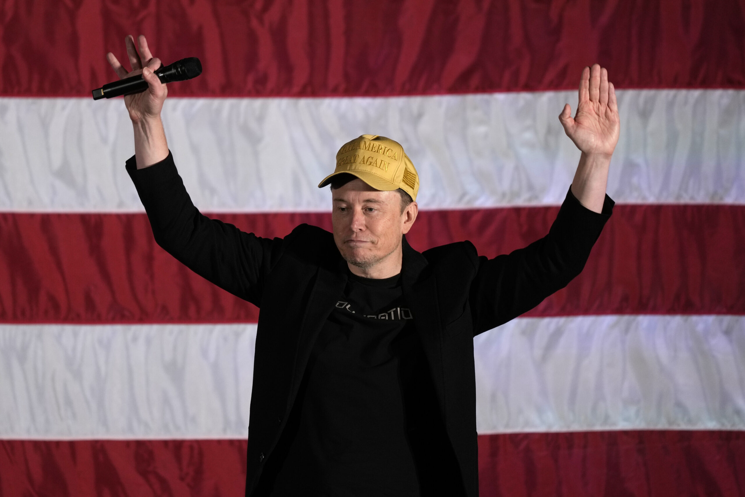 Tech-Milliardär Elon Musk steht vor einer riesigen US-amerikanischen Flagge und streckt die Hände in die Luft