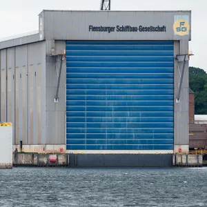 Werfthalle der Flensburger-Schiffsbau-Gesellschaft (FSG). Ein großes graues Gebäude am Wasser mit einem blauen Tor.