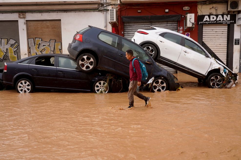 Ein Mann geht durch überflutete Straßen an Autos vorbei, die von den Wassermassen übereinander geschoben wurden.