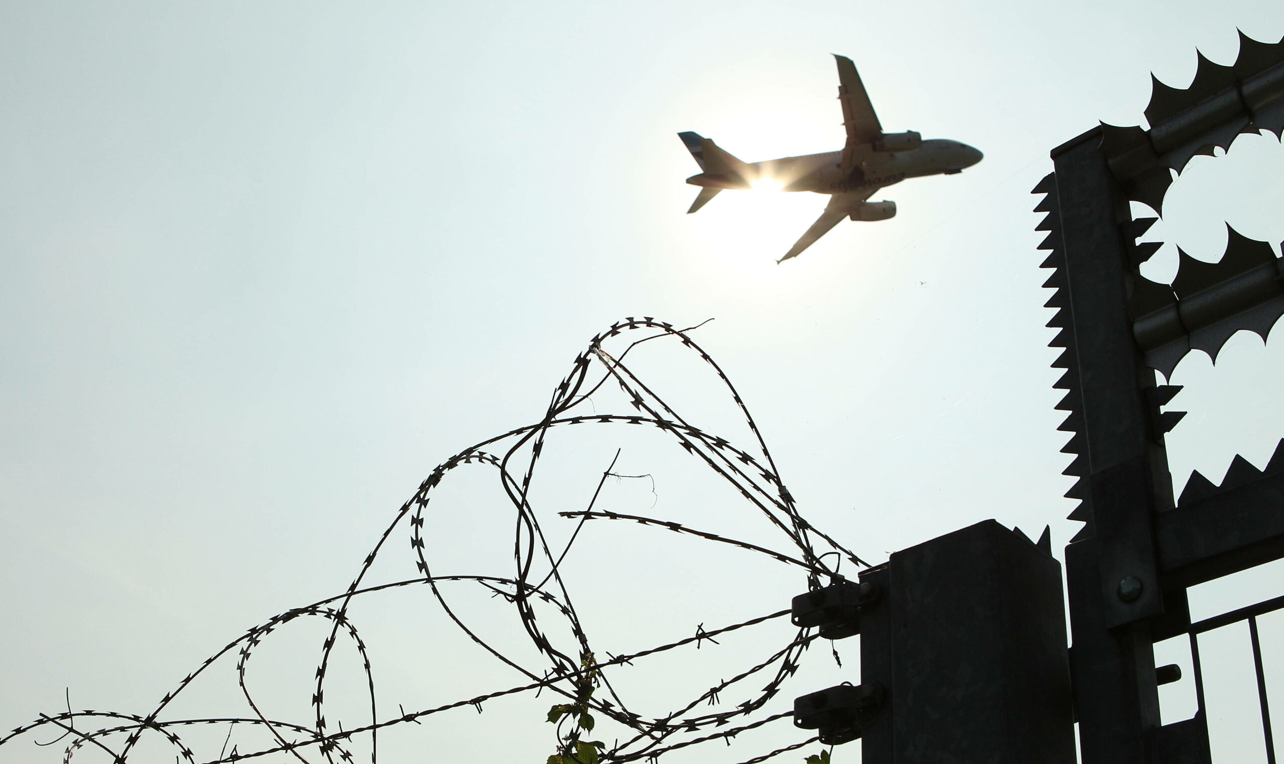 Ein Flugzeug in der Luft im Gegenlicht. Im Vordergrund ist ein hoher Stacheldrahtzaun zu sehen.