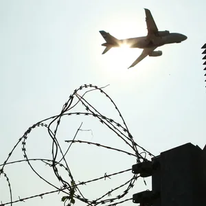 Ein Flugzeug in der Luft im Gegenlicht. Im Vordergrund ist ein hoher Stacheldrahtzaun zu sehen.