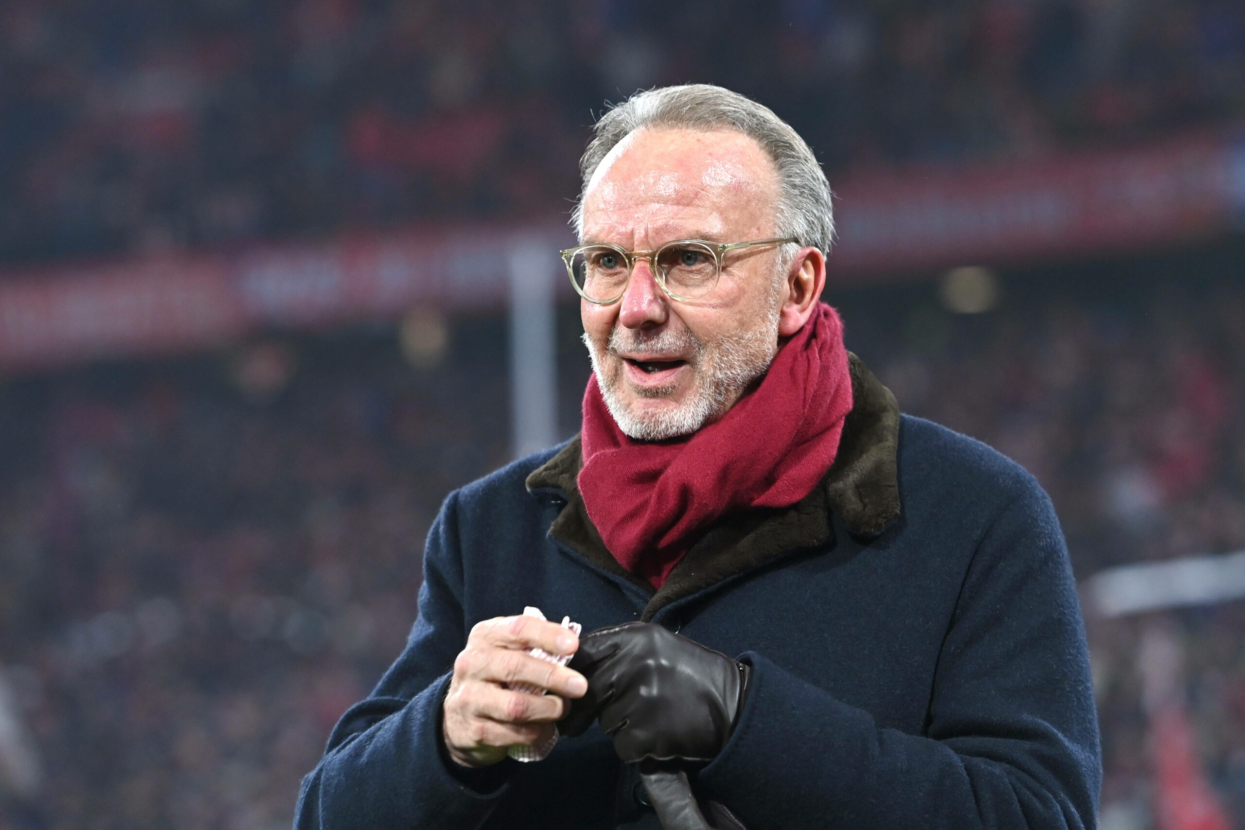 Karl-Heinz Rummenigge mit Mantel und Schal in der Allianz Arena hält ein Taschentuch in der Hand