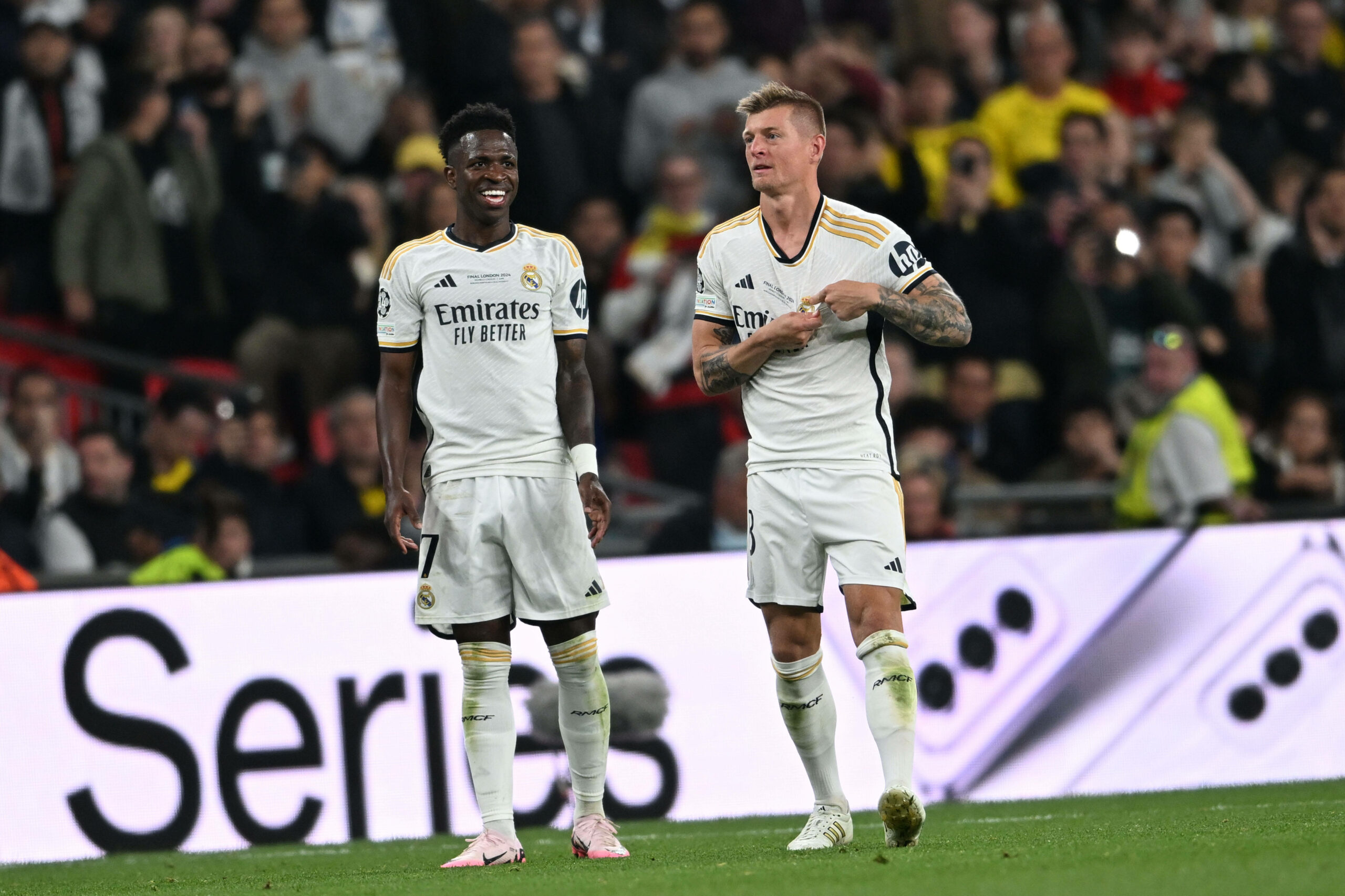 Toni Kroos zeigt auf das Wappen, während Vinicius lächelt