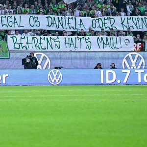Wolfsburger Fans halten ein Spruchband hoch, auf dem steht: „Egal ob Daniela oder Kevin, Behrens halt's Maul“.