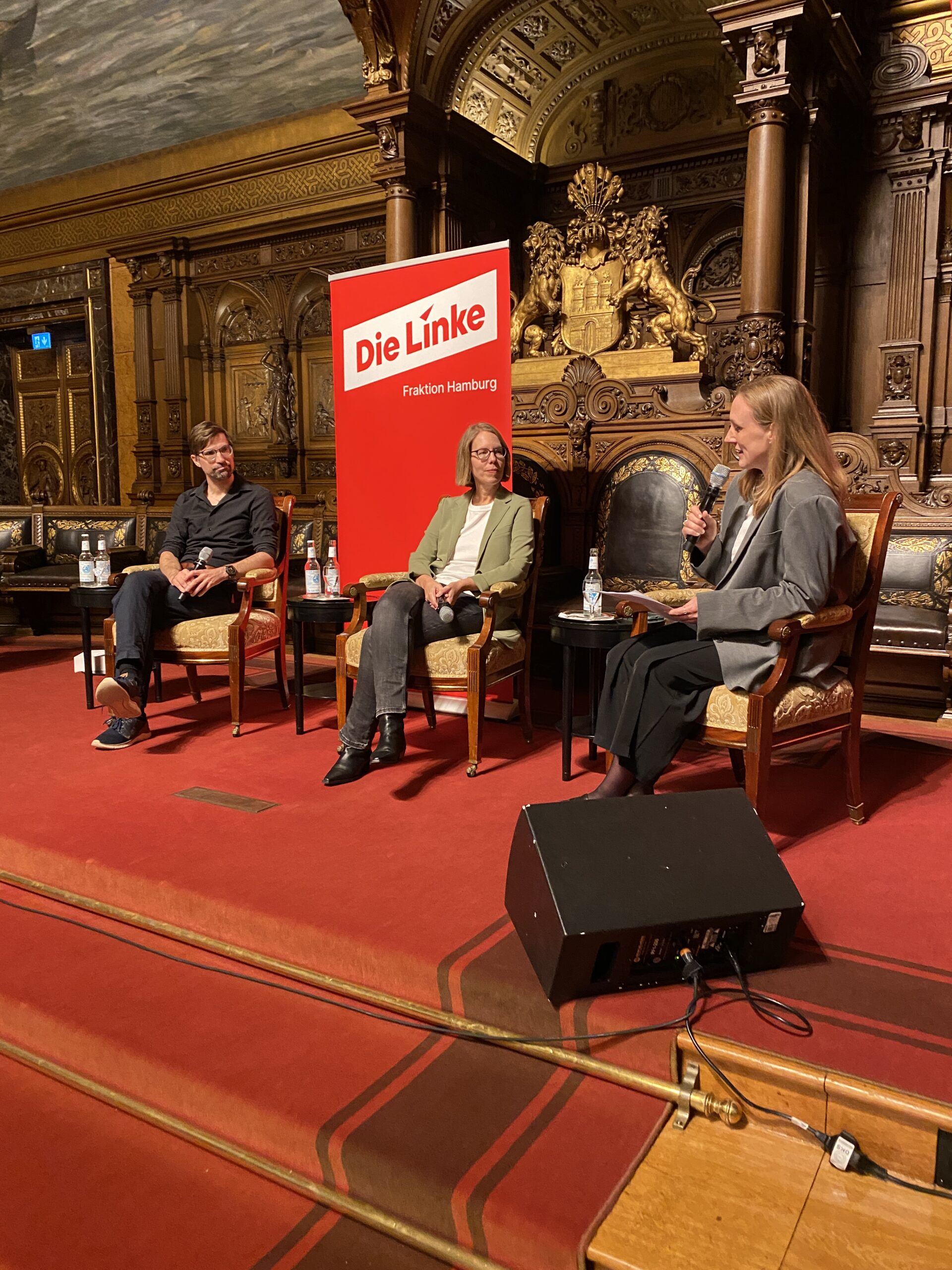 Drei Personen auf einer Bühne mit Schild „Die Linke“