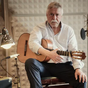 Biermann sitzt auf einem Stuhl im Studio, auf dem Schoß eine Akustikgitarre