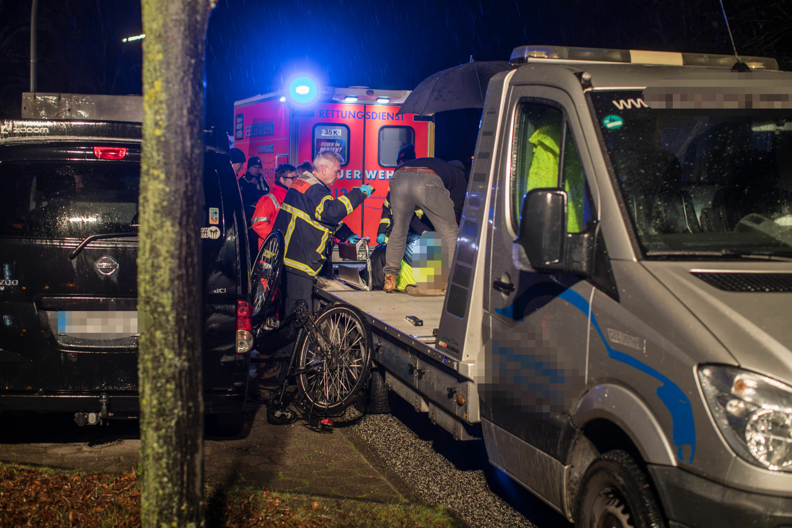 Einsatzkräfte des Rettungsdienstes stehen bei einer unkenntlicht-gemachten Person, die auf einer Transporterfläche liegt. Im Hintergrund steht ein Rettungswagen mit Blaulicht