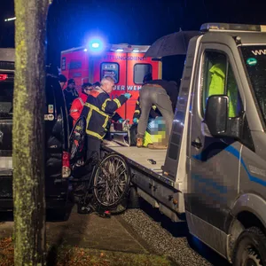 Einsatzkräfte des Rettungsdienstes stehen bei einer unkenntlicht-gemachten Person, die auf einer Transporterfläche liegt. Im Hintergrund steht ein Rettungswagen mit Blaulicht