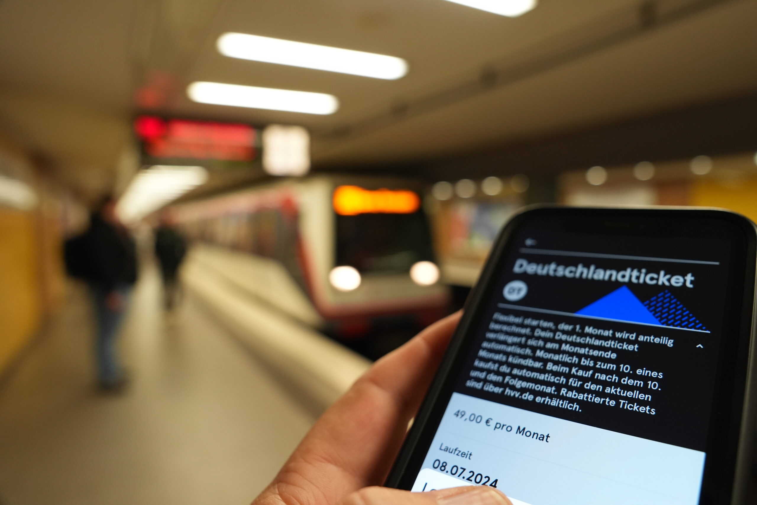 Ein Fahrgast hält ein Smartphone mit einem digitalen Deutschlandticket an einer U-Bahnstation in der Hand.