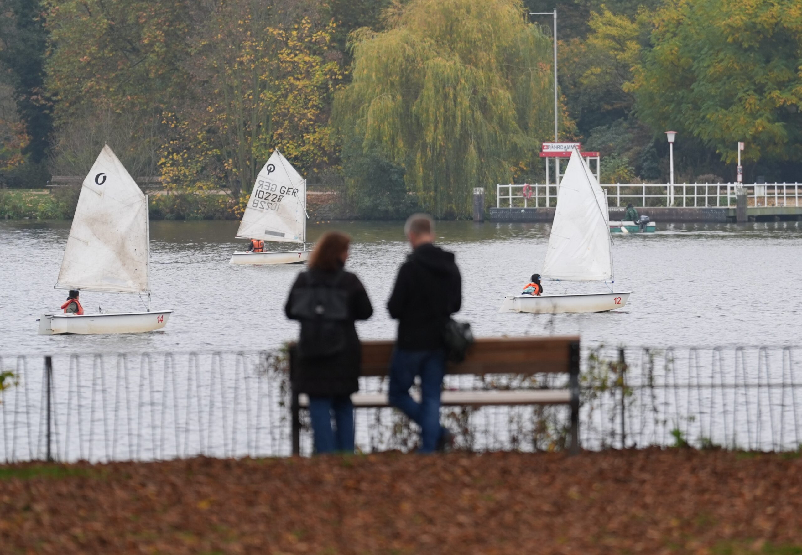 Zwei Menschen spazieren entlang der Außenalster. Im Hintergrund sind Segelboote