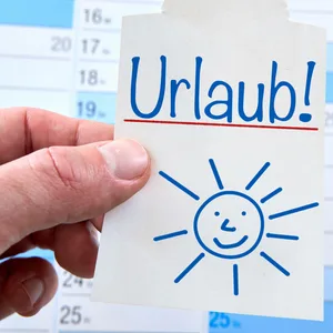 Eine Hand hält einen Zettel mit der Aufschrift „Urlaub“ vor einem Kalender.