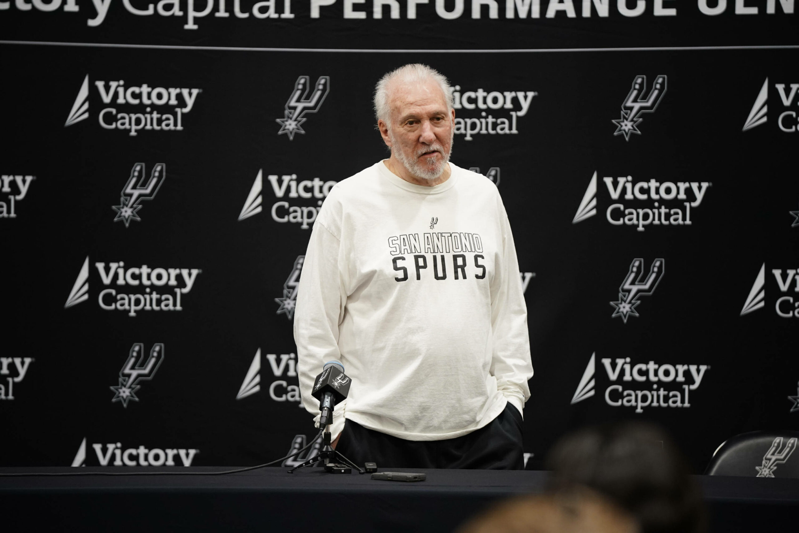 Gregg Popovich vor einer Werbetafel im Spurs Pullover