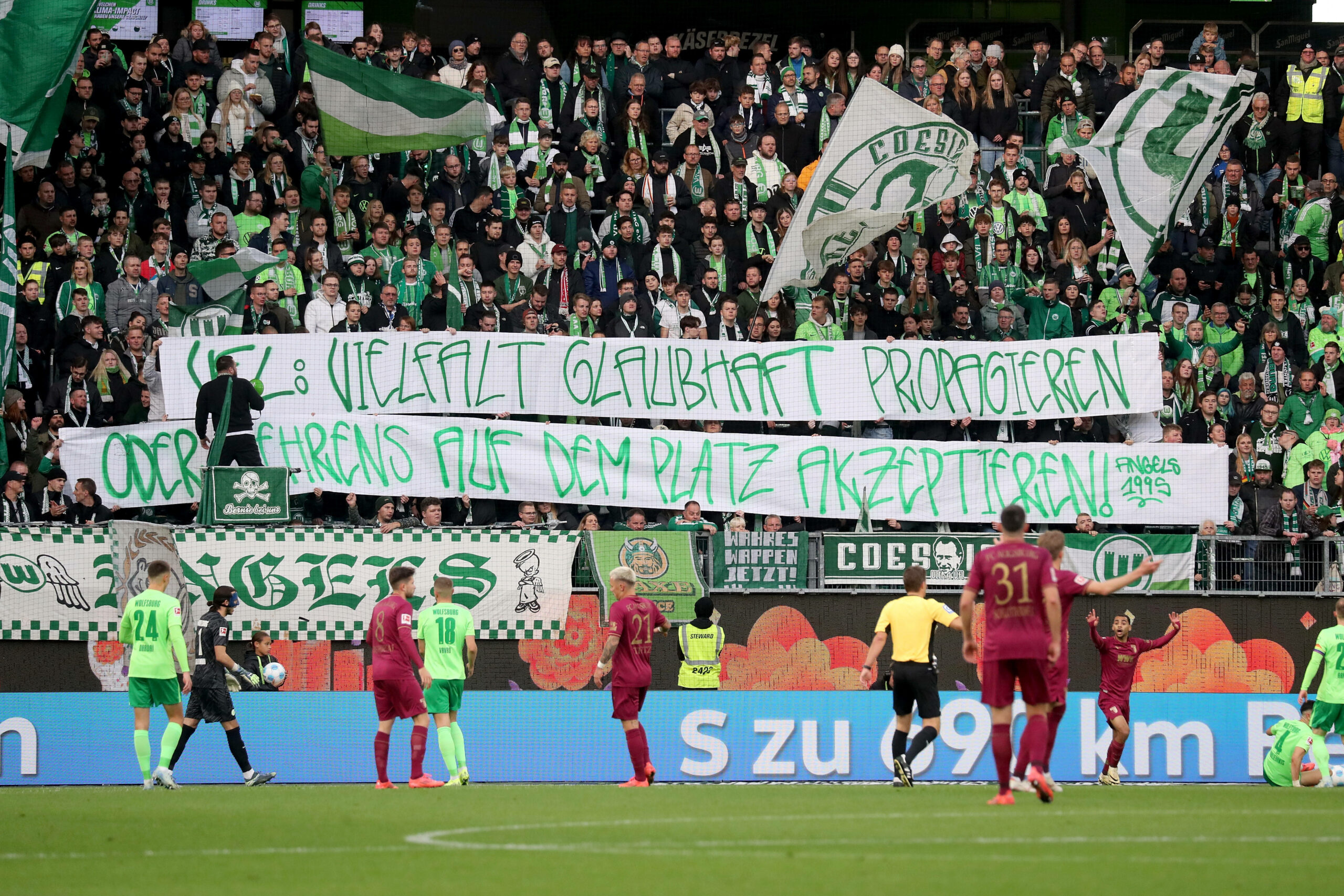 Fans des VfL Wolfsburg halten ein Transparent gegen Kevin Behrens hoch, davor stehen Spieler des VfL Wolfsburg und FC Augsburg auf dem Platz