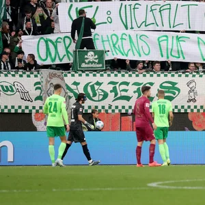 Fans des VfL Wolfsburg halten ein Transparent gegen Kevin Behrens hoch, davor stehen Spieler des VfL Wolfsburg und FC Augsburg auf dem Platz