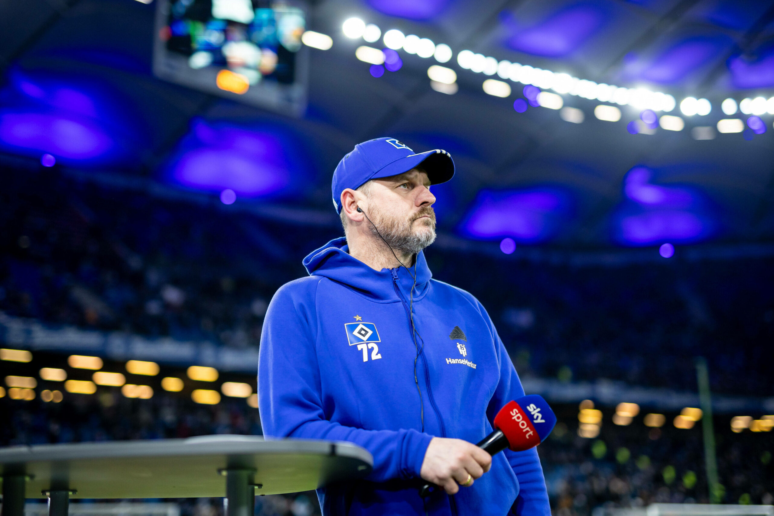 Steffen Baumgart lehnt mit Sky-Mikro auf einem Tisch am Spielfeldrand