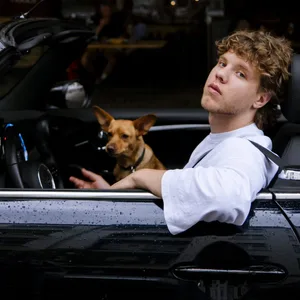 Er sitzt in einem schwarzen Auto ohne Verdeck, auf dem Beifahrersitz ein kleiner Hund