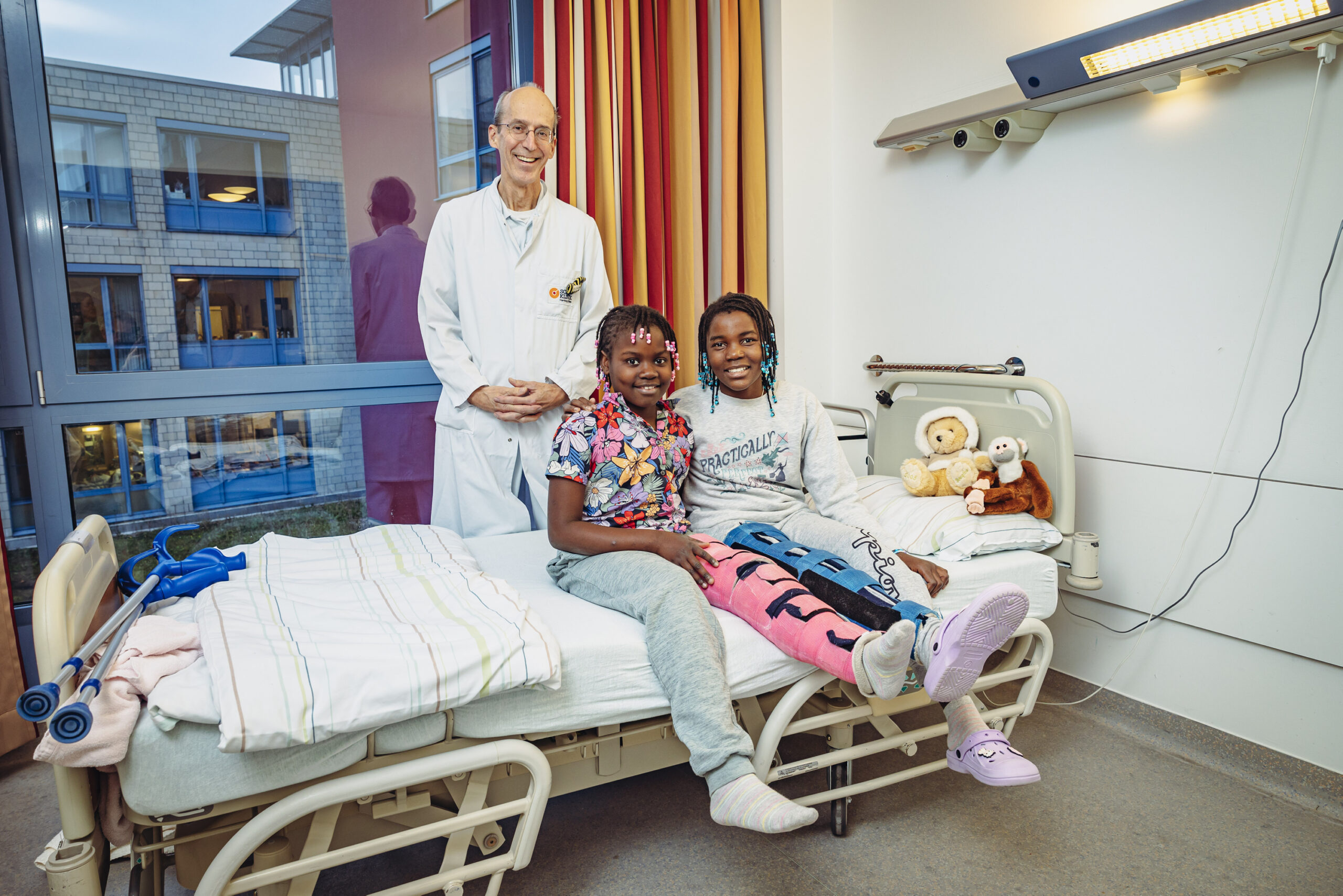 Zwei Mädchen sitzen auf einem Krankenhausbett. Hinter ihnen steht ein Arzt