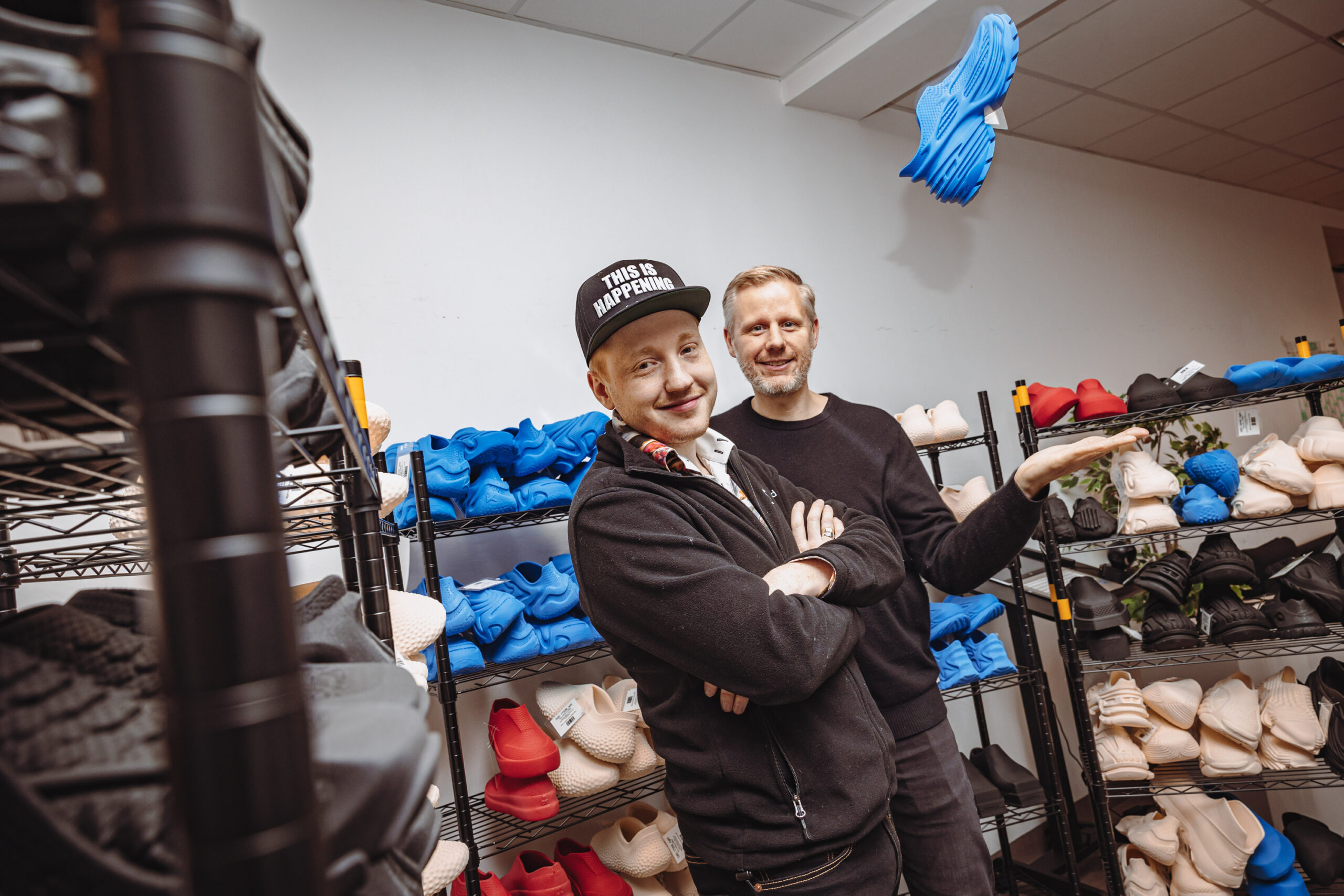 Cornelius Schmitt (links) und Michael Krause (rechts) stehen zwischen Regalen voller gedruckter Schuhe. Krause wirft einen Speaker in die Luft.