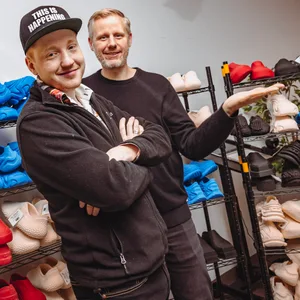 Cornelius Schmitt (links) und Michael Krause (rechts) stehen zwischen Regalen voller gedruckter Schuhe. Krause wirft einen Speaker in die Luft.