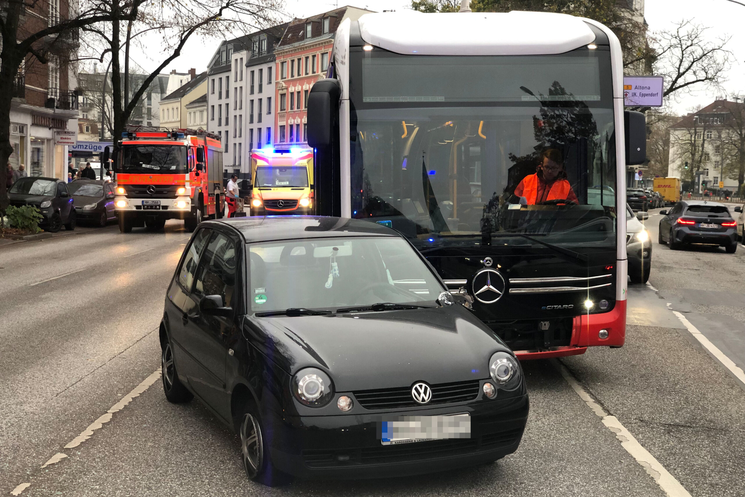 Ein PKW steht mit leicht beschädigtem Außenspiegel vor einem Bus. Im Hintergrund stehen Feuerwehr und Rettungskräfte