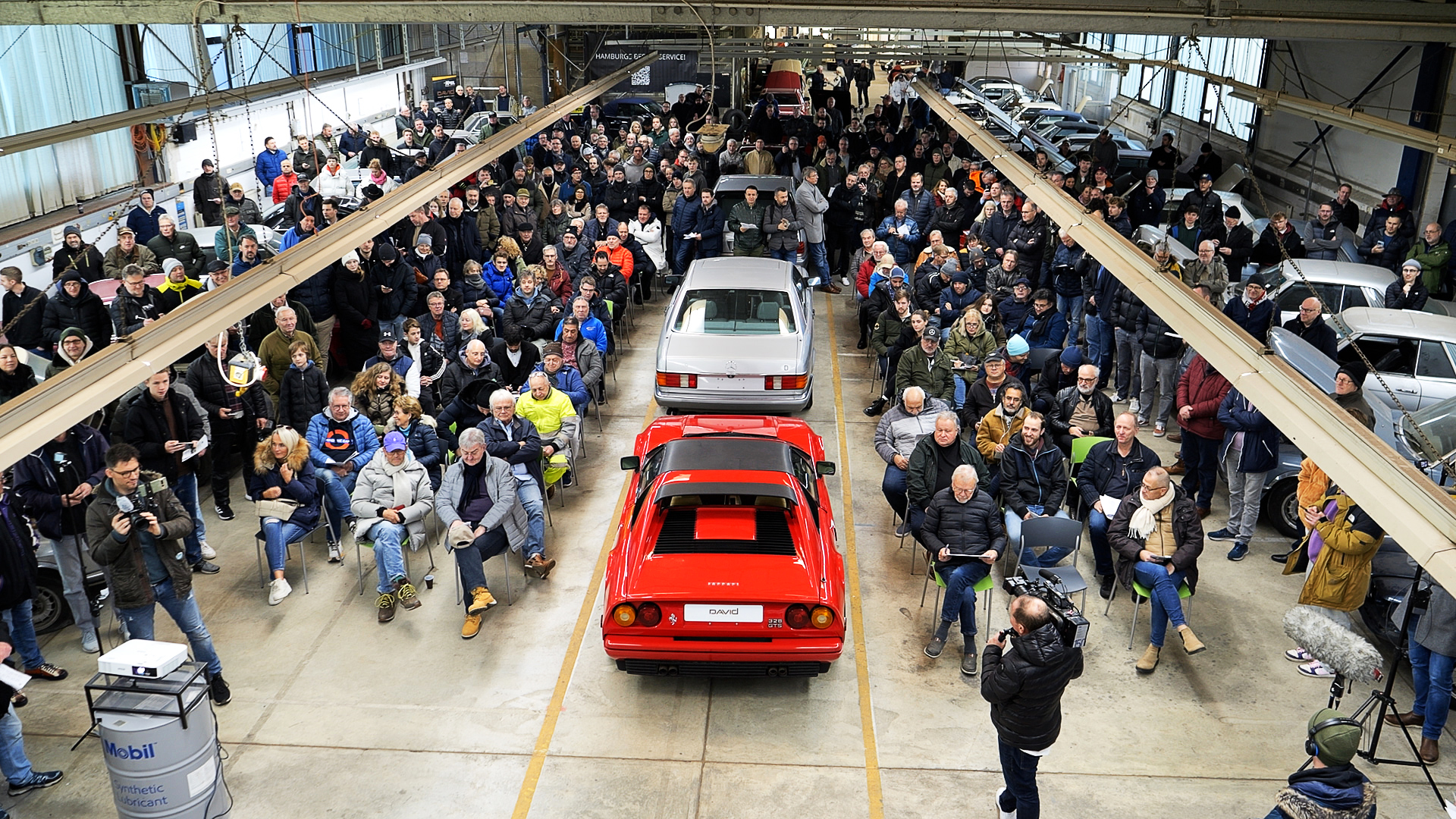 Zahlreiche Menschen sitzen in einer Halle für die Versteigerung der Oldtimer.