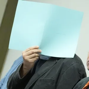 Ein Mann verdeckt sein Gesicht im Gericht mit einem Blatt Papier. Neben ihm steht sein Verteidiger