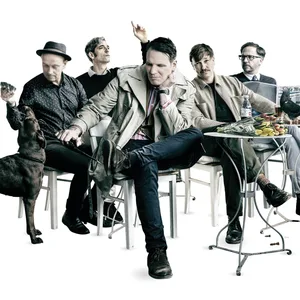 Die Band sitzt an Café-Tischen, einer liest Zeitung, links ist ein Hund zu sehen.