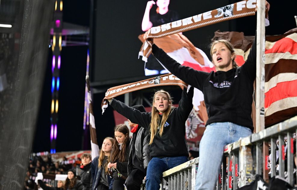 Fans des FC St. Pauli sitzen auf dem Zaun der Tribüne, halten ihre Schals in die Höhe und singen