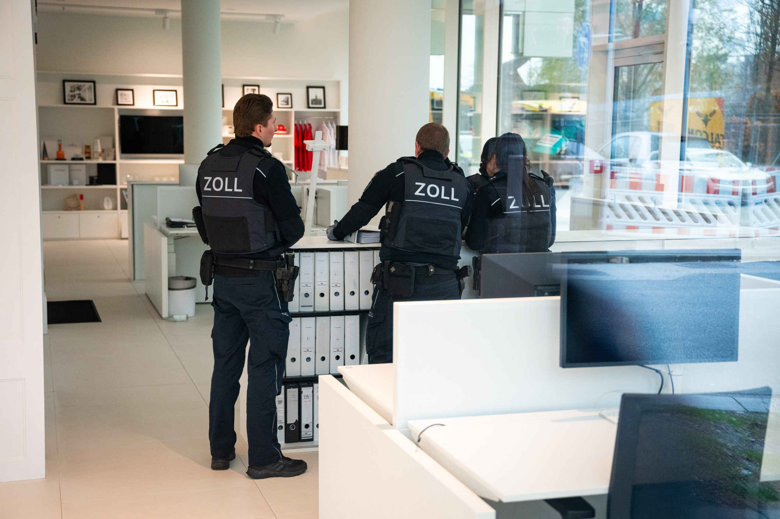 Angestellte des Zoll durchsuchen ein Bürogebäude. Zu Dritt stehen sie vor einem Schrank voller Ordner.