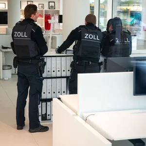 Angestellte des Zoll durchsuchen ein Bürogebäude. Zu Dritt stehen sie vor einem Schrank voller Ordner.