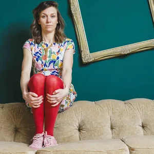 Eine Frau sitzt auf einem Sofa, im Hintergrund hängt ein schiefer Bilderrahmen.