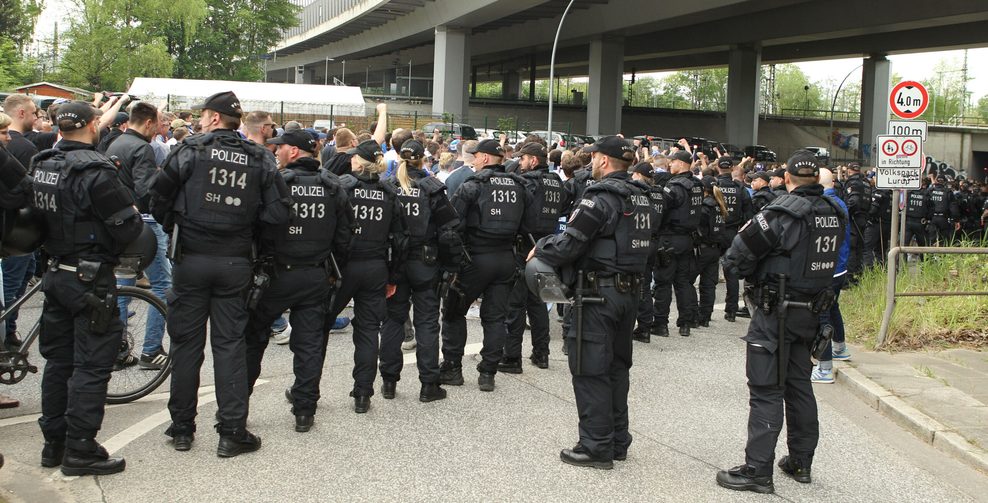 Im Vordergrund sind Polizisten zu sehen. Im Hintergrund Fans die unter einer Brücke durchlaufen