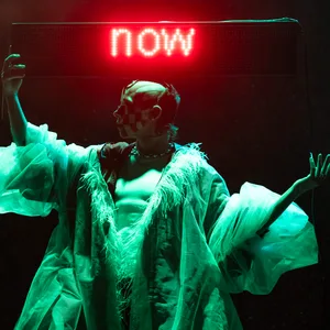 Drag-Star Wet Mess hält ein Neonschild mit der Aufschrift „now“ über dem Kopf.