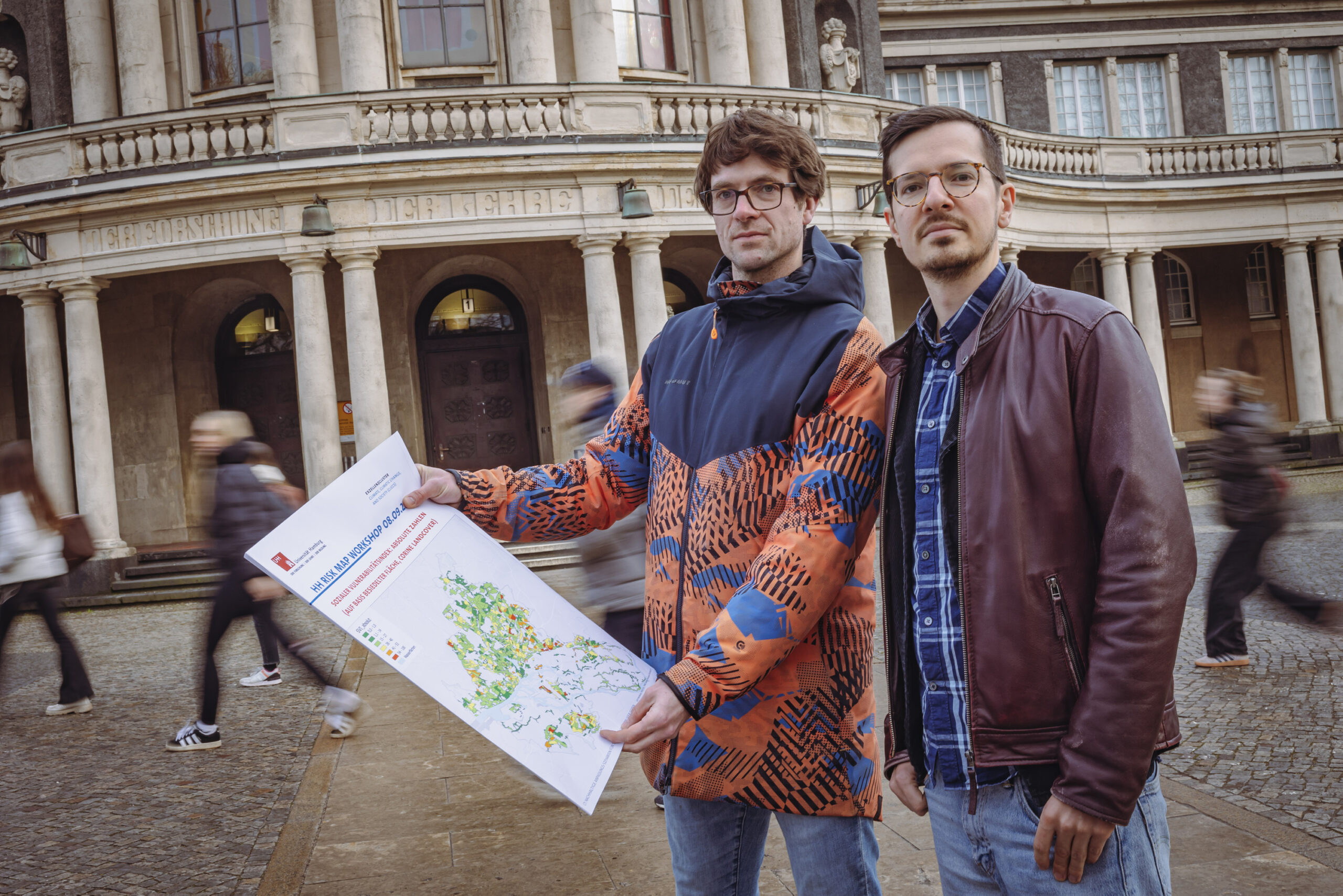 Die zwei Klimaforscher stehen vor dem Hauptgebäude der Universität Hamburg. Sie halten eine Karte die die Auswirkungen des Klimawandels auf soziale Gruppen zeigt.