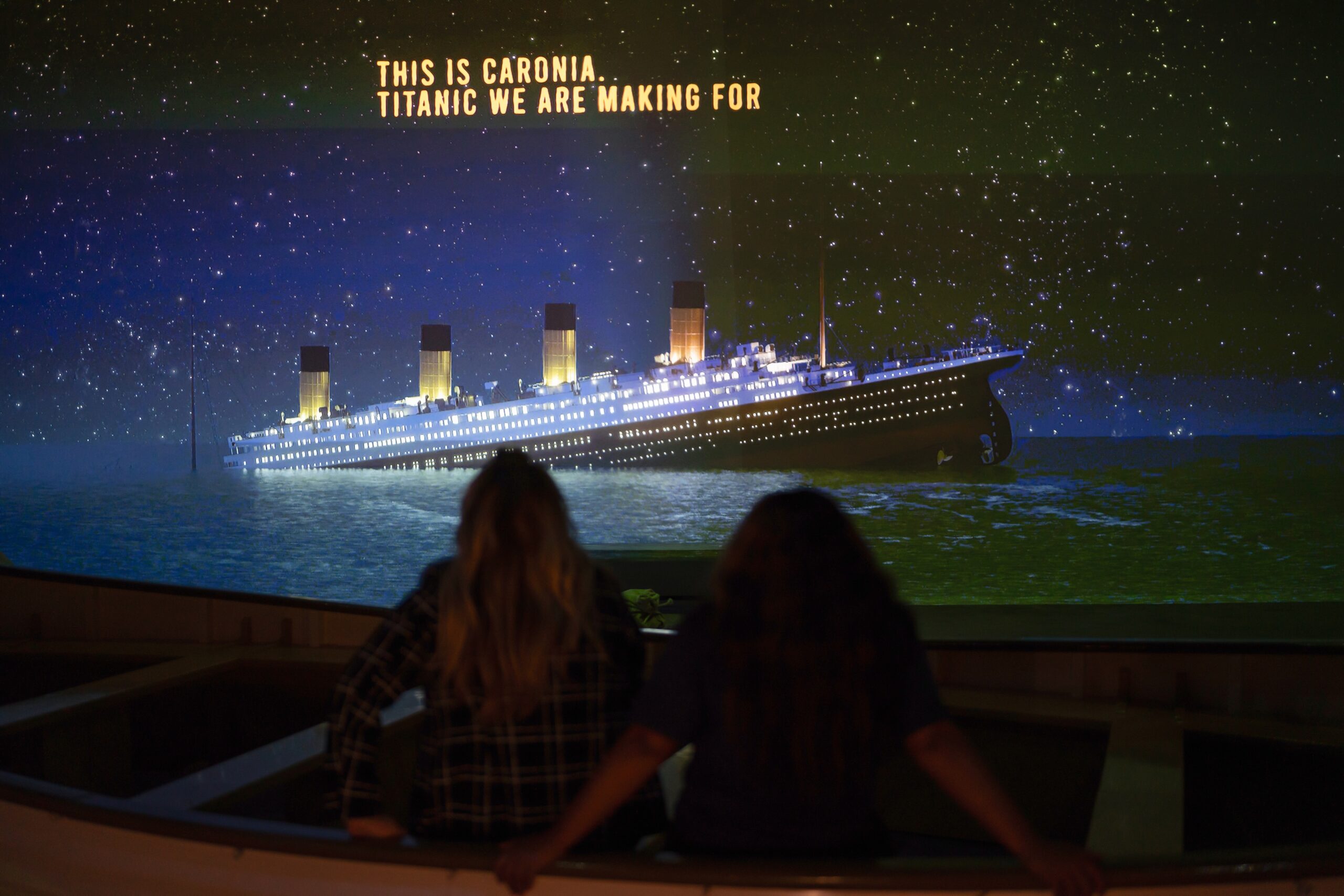 Zwei Menschen schauen auf eine Leinwand, man sieht dort die „Titanic“ untergehen