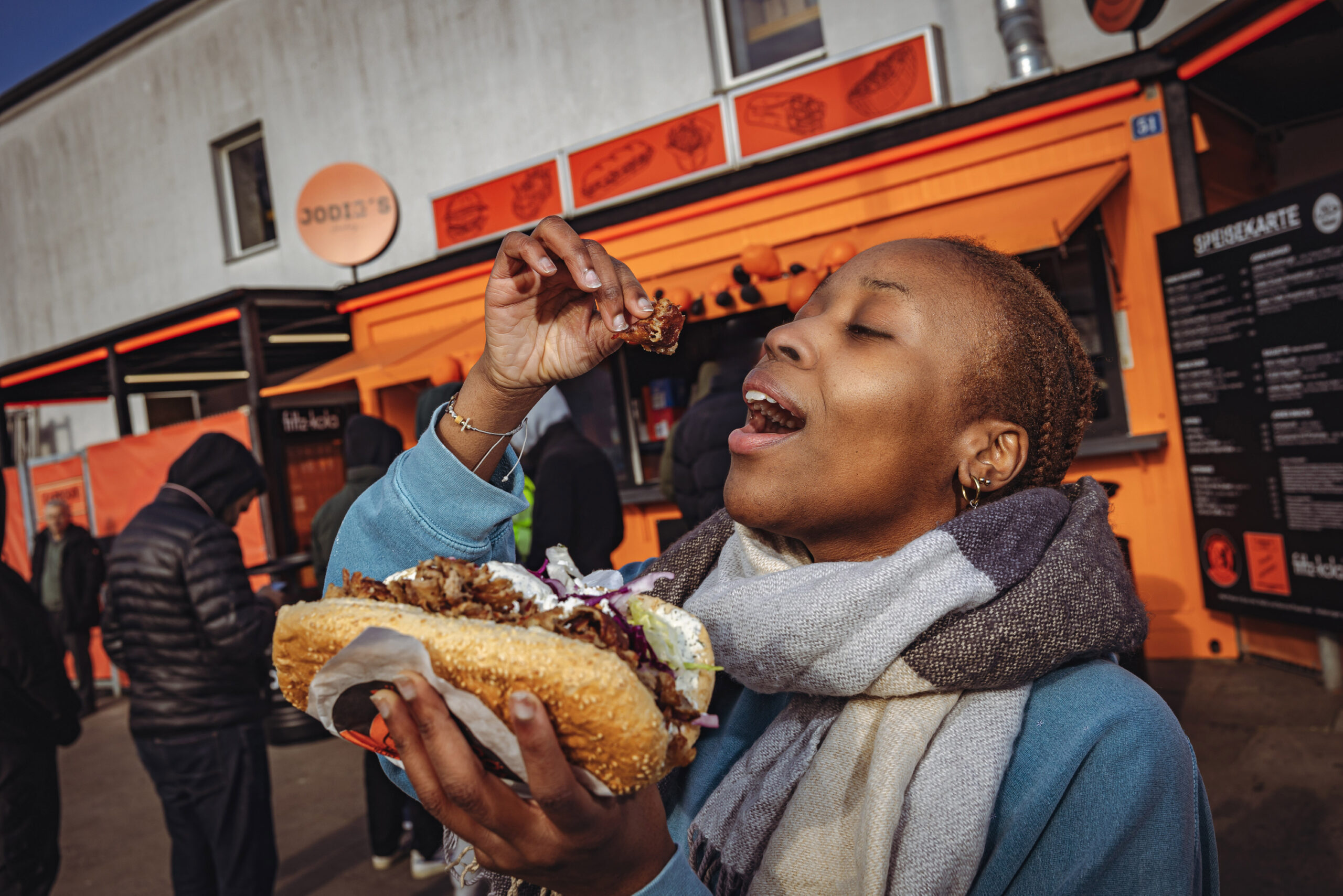 Eine Frau hält in der linken Hand einen Döner, in der Rechten, ein Stück Fleisch vor einem Dönerladen