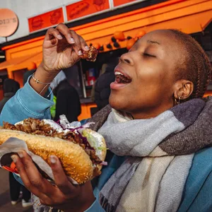 Eine Frau hält in der linken Hand einen Döner, in der Rechten, ein Stück Fleisch vor einem Dönerladen