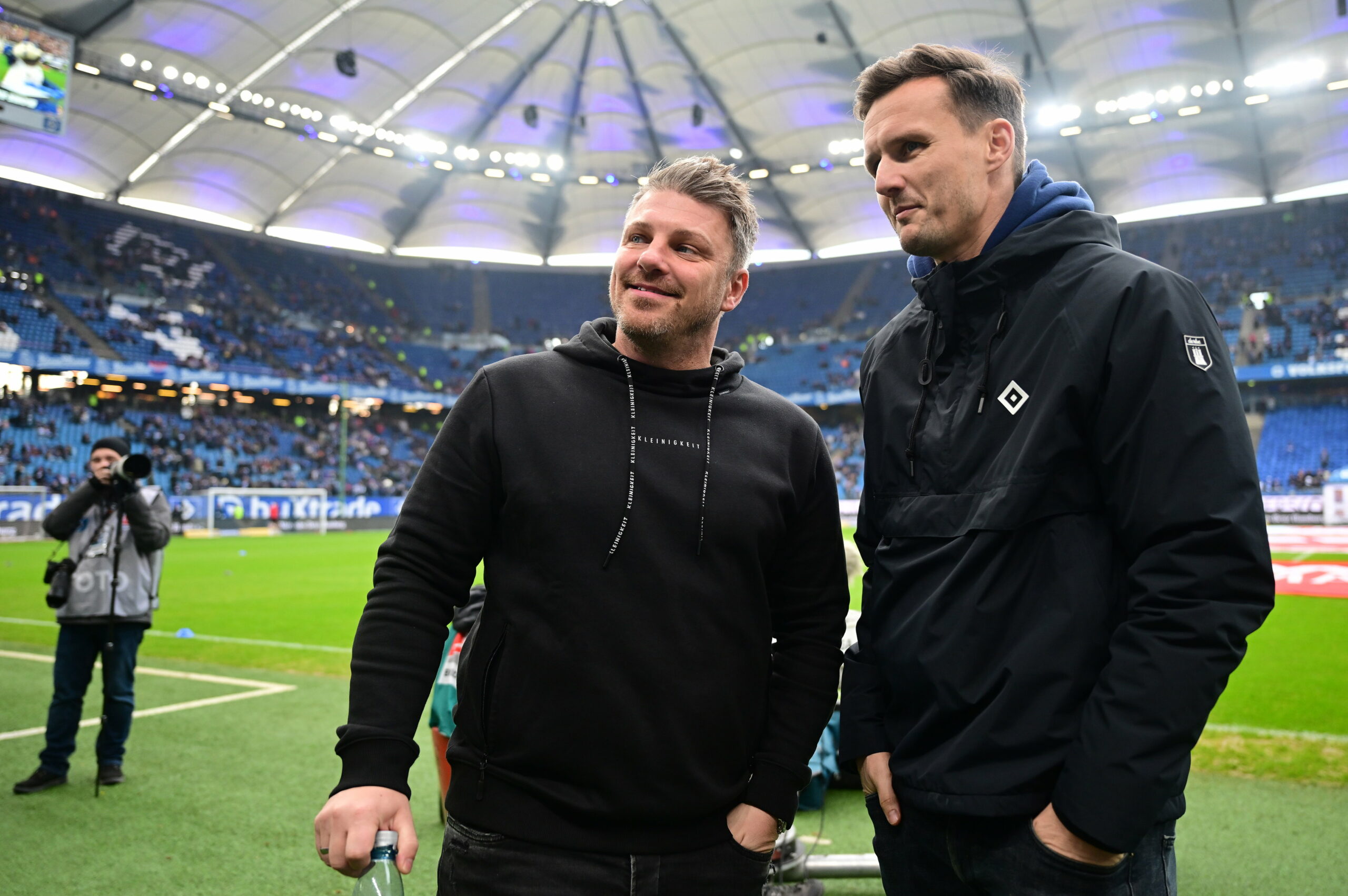 Lukas Kwasniok und Claus Costa lachen am Spielfeldrand im Volksparkstadion in eine Kamera