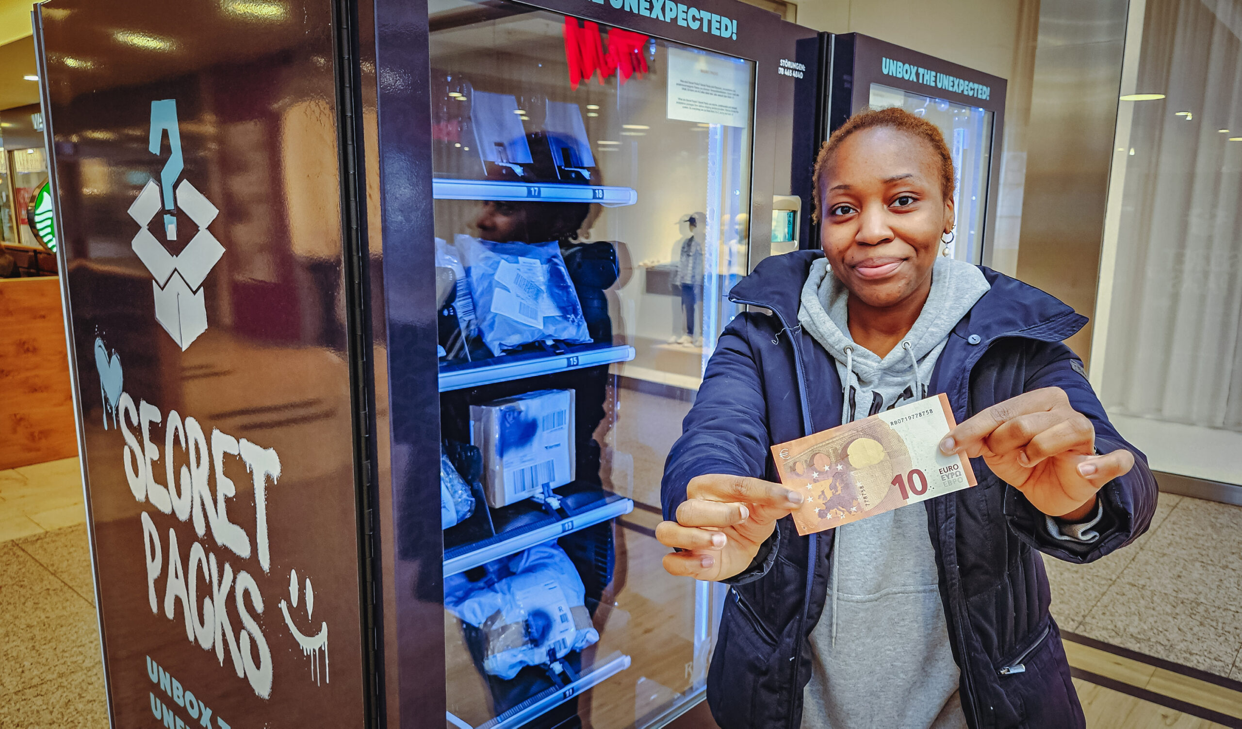 Die MOPO Reporterin Naomi Wita steht vor einem Mystery Box Automaten und hält einen zehn Euro Schein in die Kamera