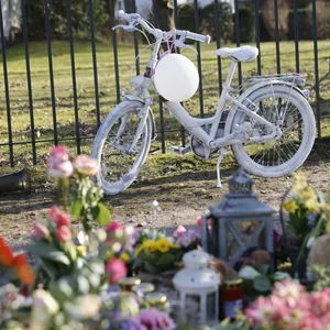 Ein weiß lackiertes Kinderfahrrad steht vor einem Zaun am Unfallort. Davor liegen Blumen.