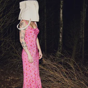 Eine Frau steht in Kleid und mit einer Tasche über den Kopf gestülpt im Wald.