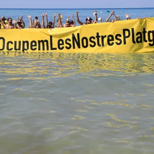 Viele Menschen stehen am Strand und halten ein Plakat hoch, auf dem steht ”Besetzen wir (selbst) unsere Strände“