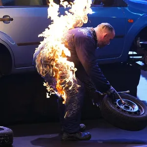 Ein Stuntman wechselt Reifen an einem Auto, während sein Anzug in Flammen steht.
